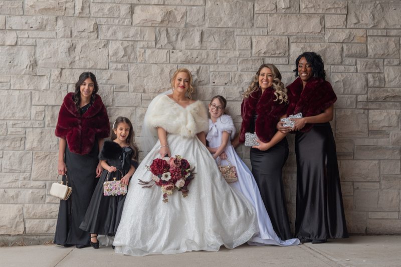 A bridal party poses joyfully against a stone wall, the bride in white with a burgundy bouquet, bridesmaids in black gowns.