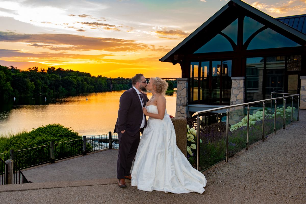A bride and groom kiss on a waterfront terrace at sunset, surrounded by a modern building and greenery.