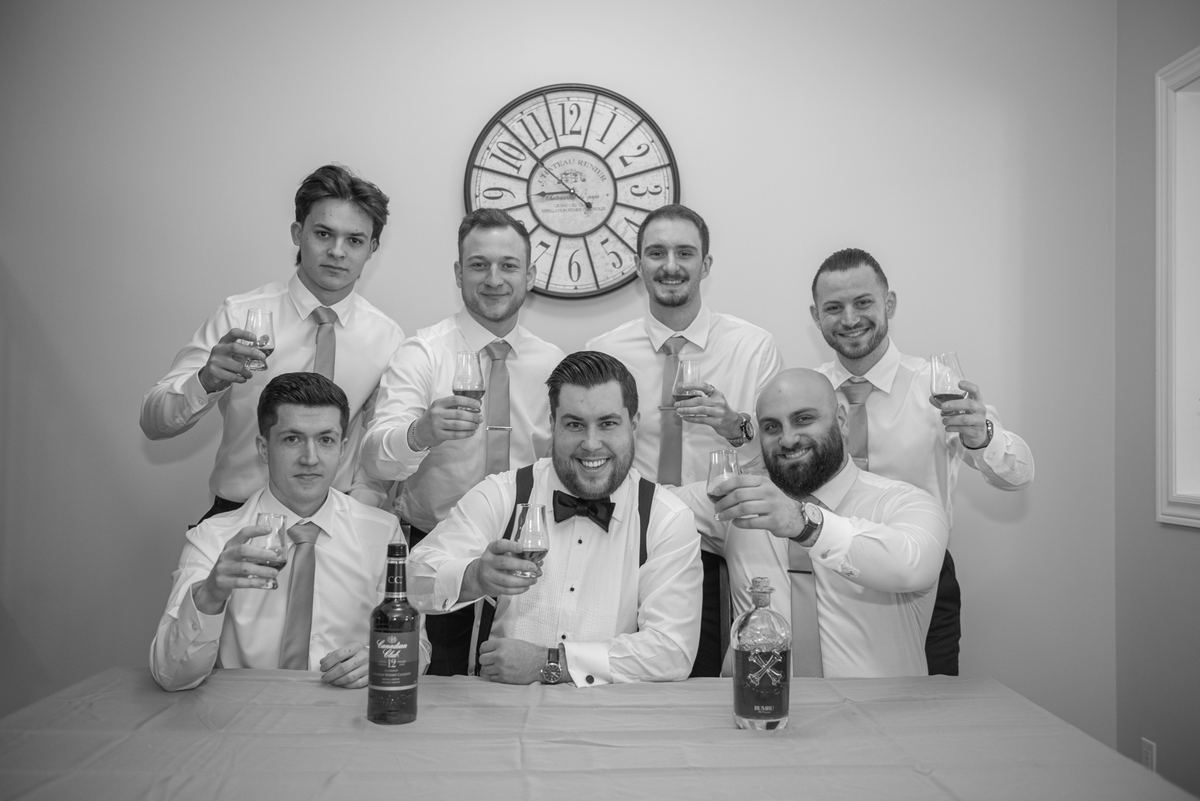 Seven groomsmen raise glasses in a toast around a table with bottles and a decorative clock behind them, celebrating.