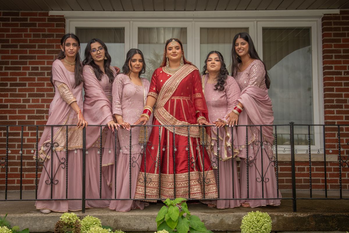 A bride in red and gold stands with five mauve-clad bridesmaids on a balcony against a brick building.