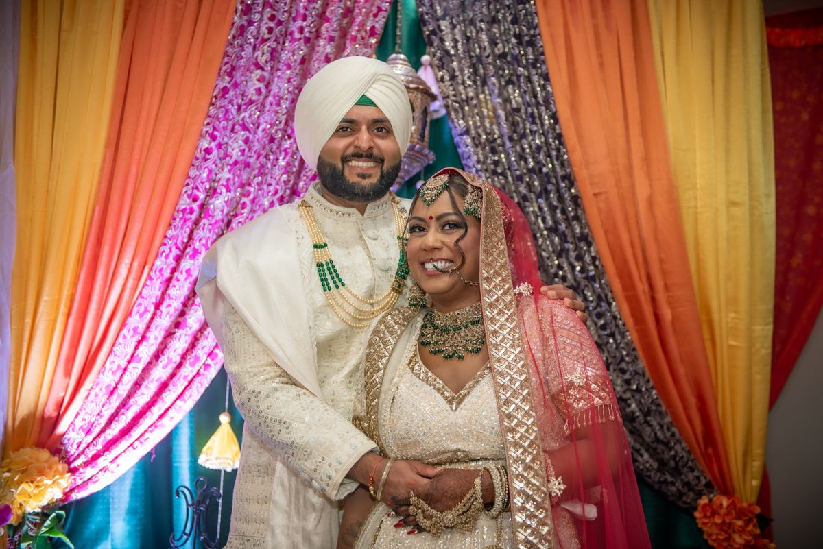 A smiling South Asian bride and groom pose in traditional attire against colorful drapes, exuding joy.
