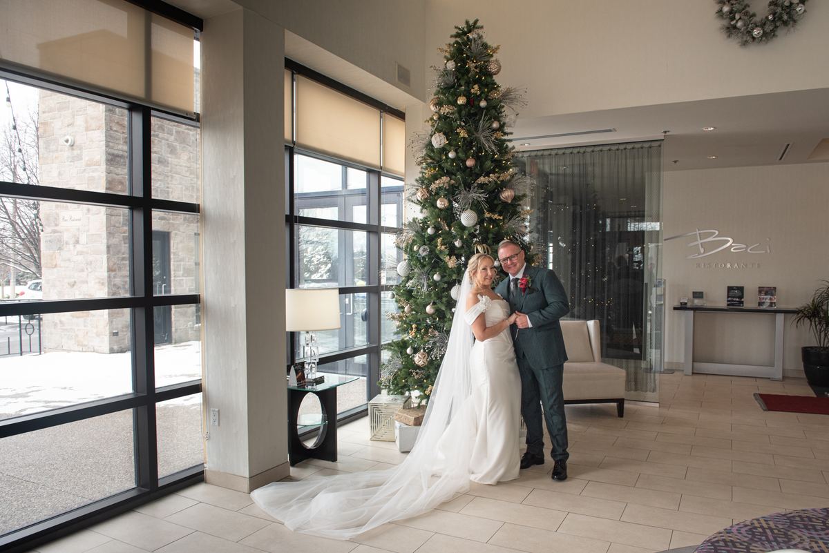 A bride and groom stand in a modern lobby beside an ornate Christmas tree, smiling as natural light streams through windows.
