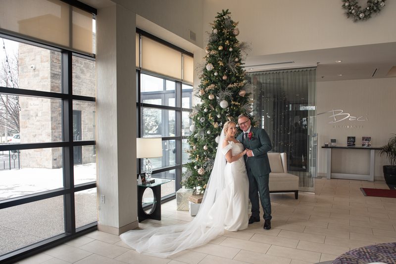 A bride and groom stand in a modern lobby beside an ornate Christmas tree, smiling as natural light streams through windows.