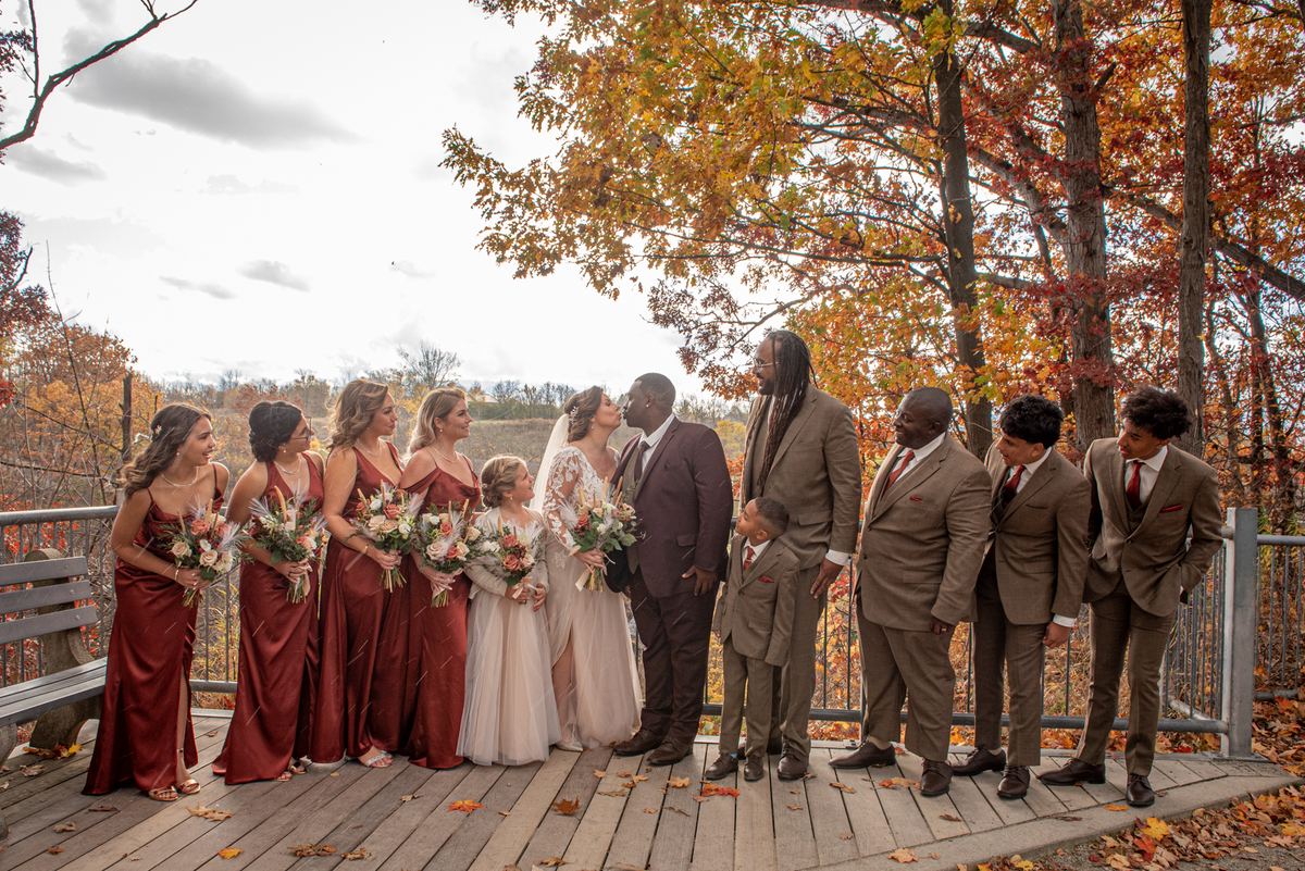 Bride and groom kiss on a wooden deck surrounded by smiling bridesmaids in burgundy gowns and groomsmen in brown suits.