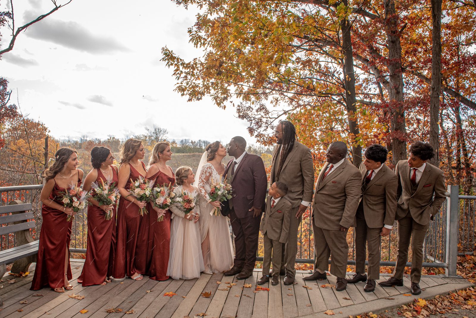 Bride and groom kiss on a wooden deck surrounded by smiling bridesmaids in burgundy gowns and groomsmen in brown suits.