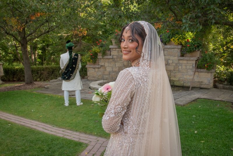 A smiling bride in an ornate outfit and veil turns to face the camera beside her groom in traditional attire.