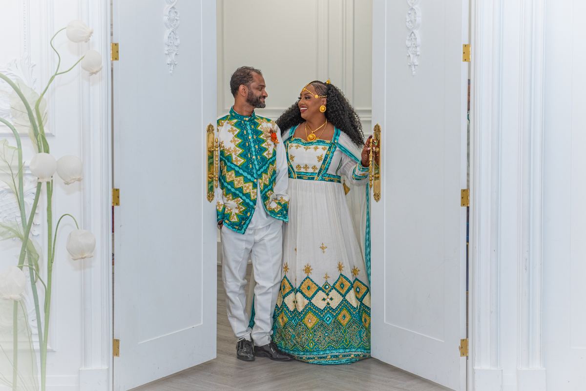 Salem and Shannon stand smiling at each other between ornate white doors, framed by decorative floral arrangements.