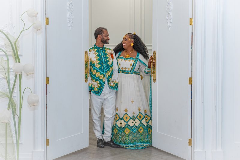 Salem and Shannon stand smiling at each other between ornate white doors, framed by decorative floral arrangements.