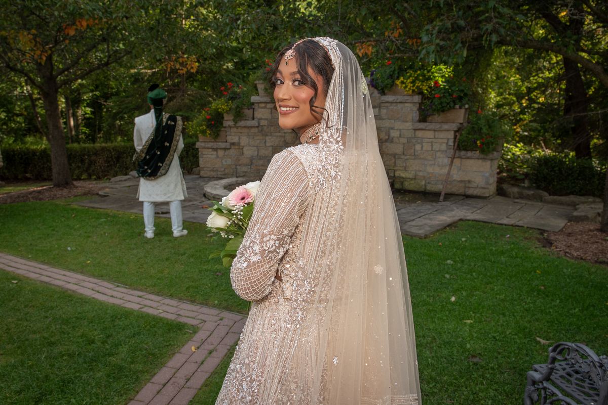 A smiling bride in a cream lehenga and veil holds flowers as the groom stands on a garden path, both bathed in soft daylight.