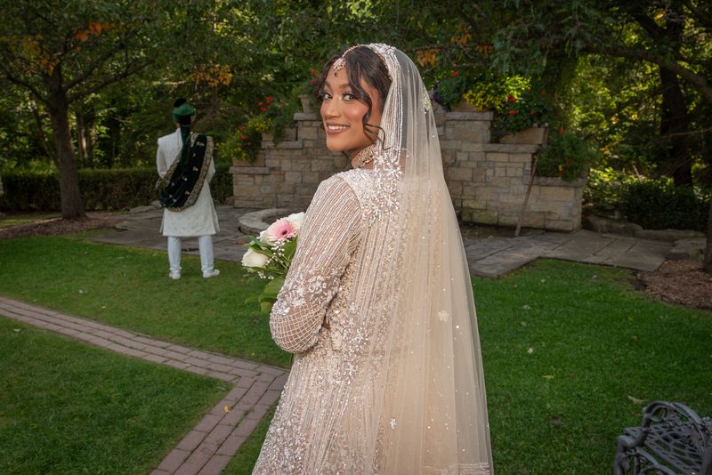 A smiling bride in a cream lehenga and veil holds flowers as the groom stands on a garden path, both bathed in soft daylight.