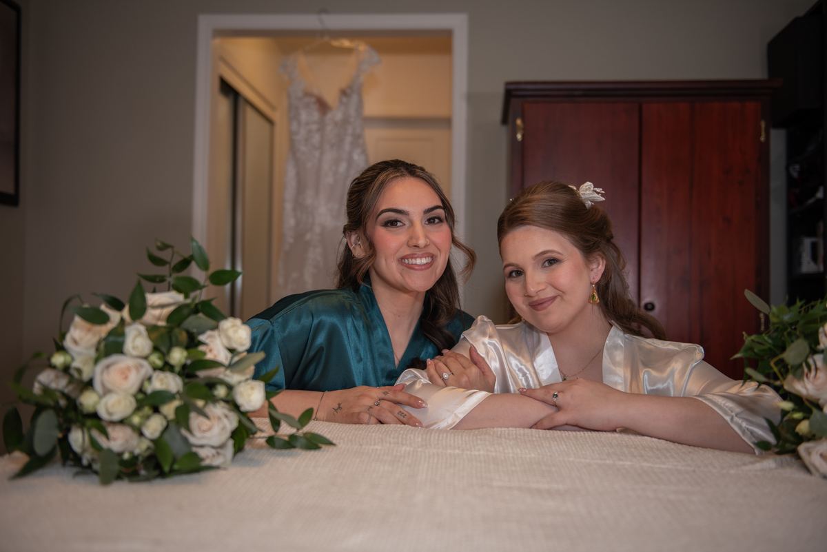Two brides smile warmly on a bed in a softly lit bridal suite with a wedding gown and floral arrangements nearby.