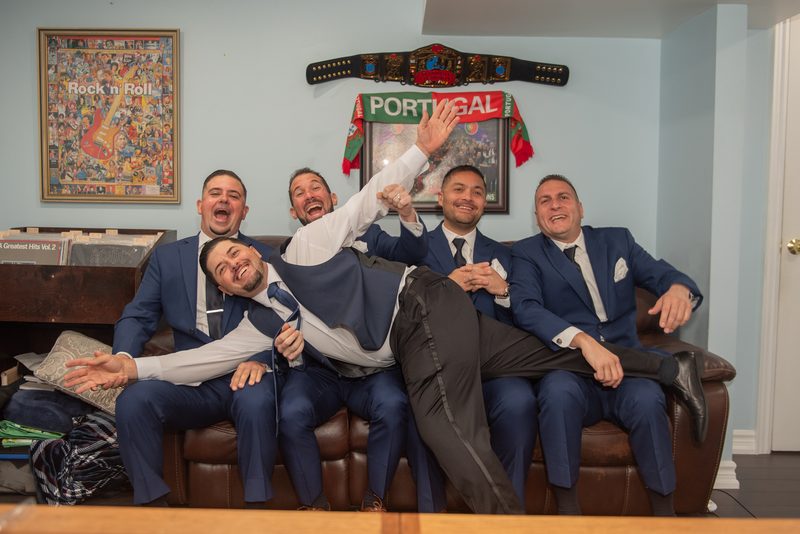 Five groomsmen in blue suits laugh and pose playfully on a leather couch, surrounded by personal memorabilia.