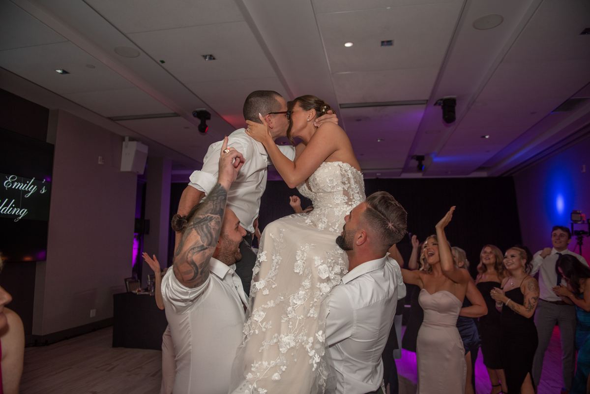The bride and groom kiss while being lifted by two groomsmen amid cheering guests under colorful stage lights.
