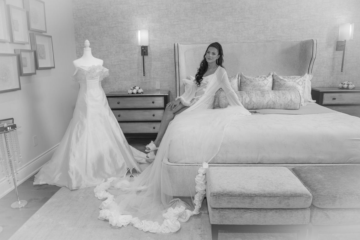 A bride sits gracefully on a large bed in an elegant bedroom, gazing toward the camera with a serene expression.