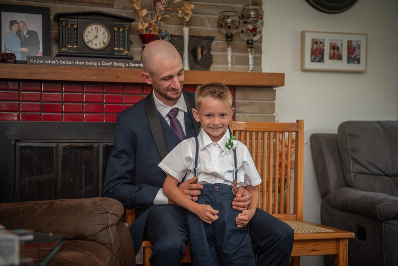 A man in a suit sits with a young boy dressed formally by the fireplace, surrounded by family photos.