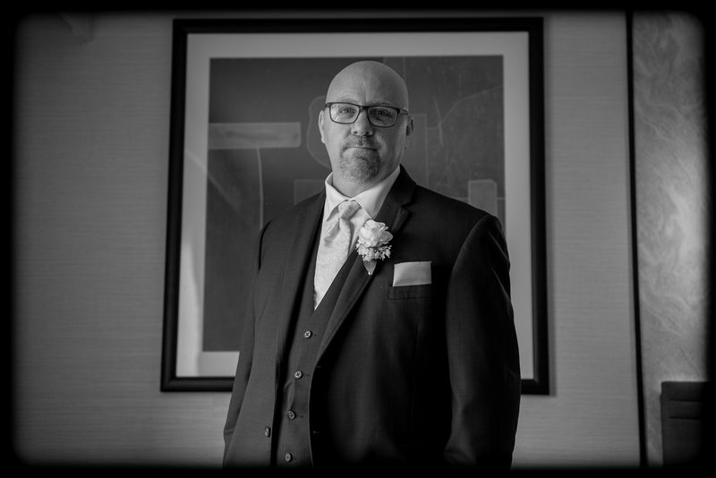A formally dressed bald man with glasses and a boutonnière stands for a portrait against a minimalist background.