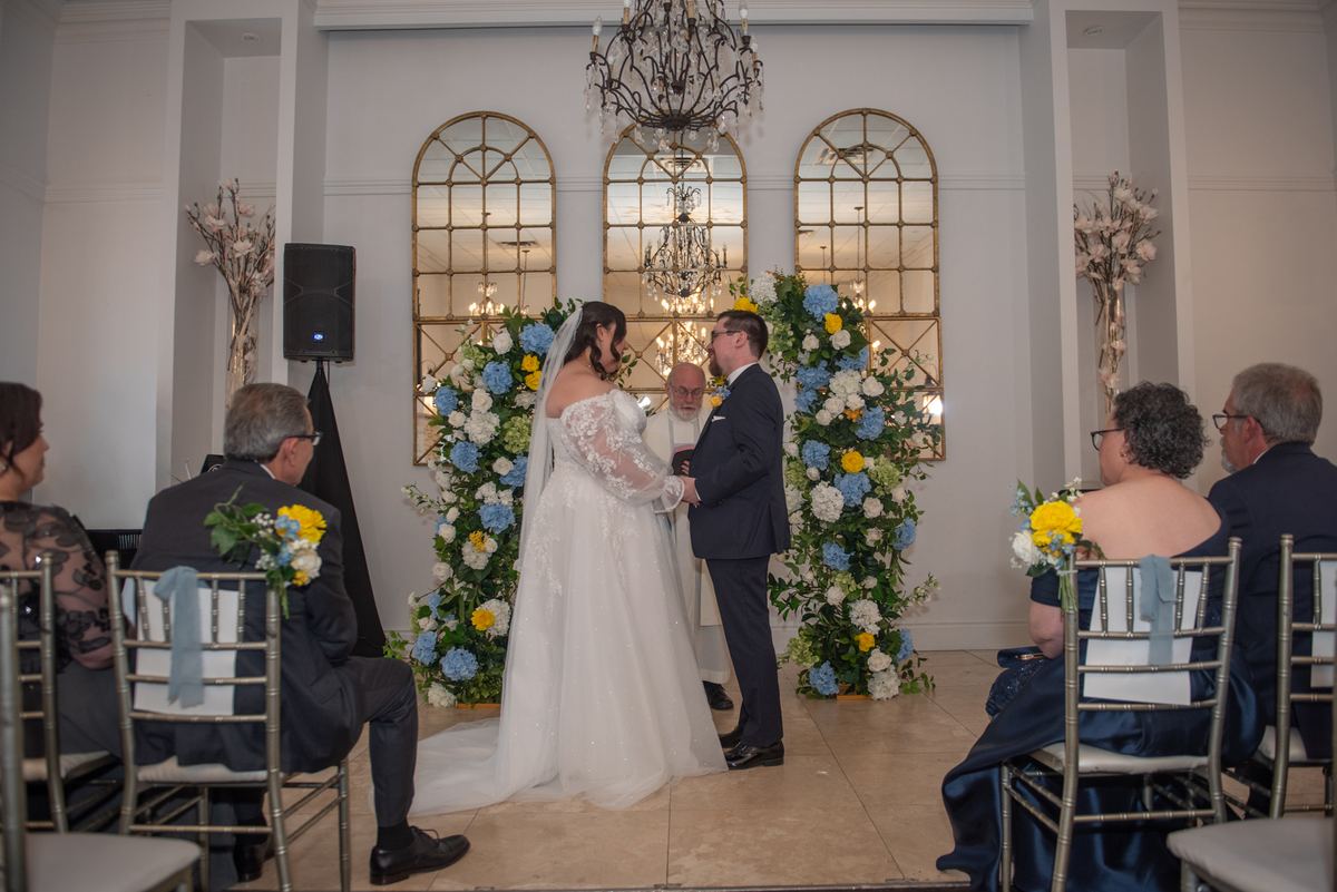 A bride and groom exchange vows at an altar under a chandelier, surrounded by guests in rows of chairs.