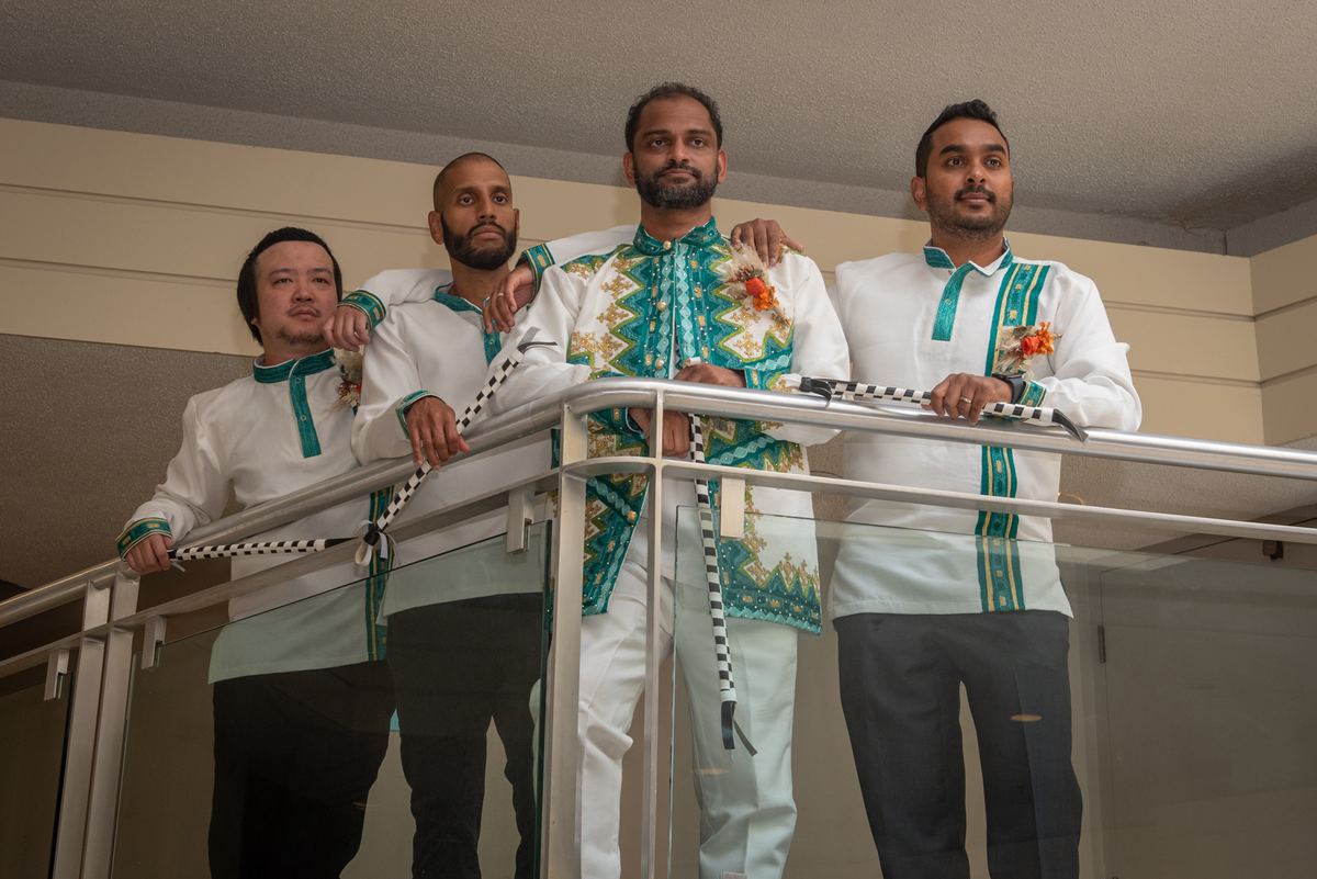 Four men in white and teal attire stand on a glass balcony, facing the camera with composed expressions.