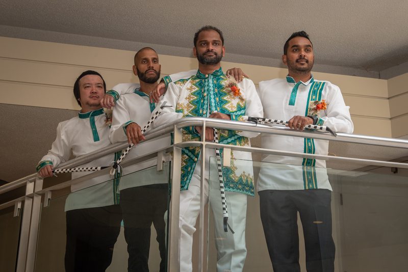 Four men in white and teal attire stand on a glass balcony, facing the camera with composed expressions.
