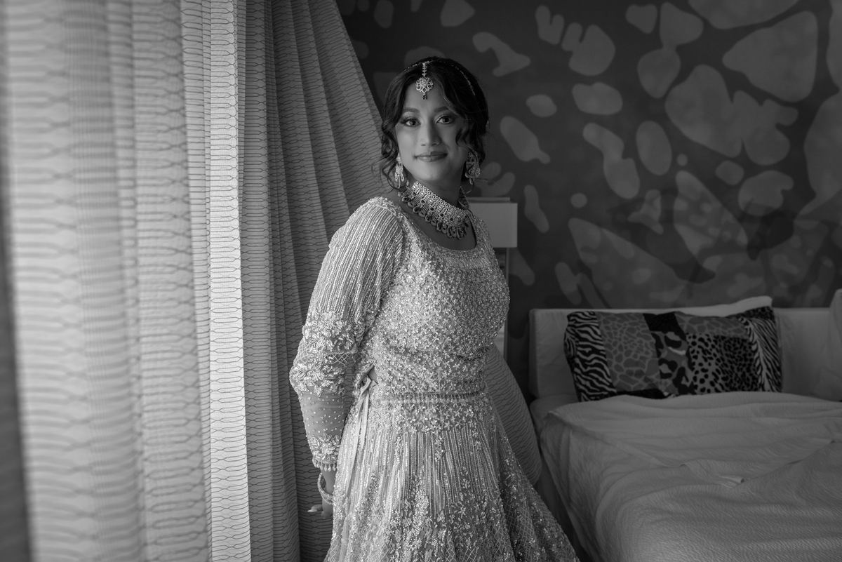 A bride in an ornate traditional gown and jewelry stands calmly in a hotel room with light filtering through curtains.