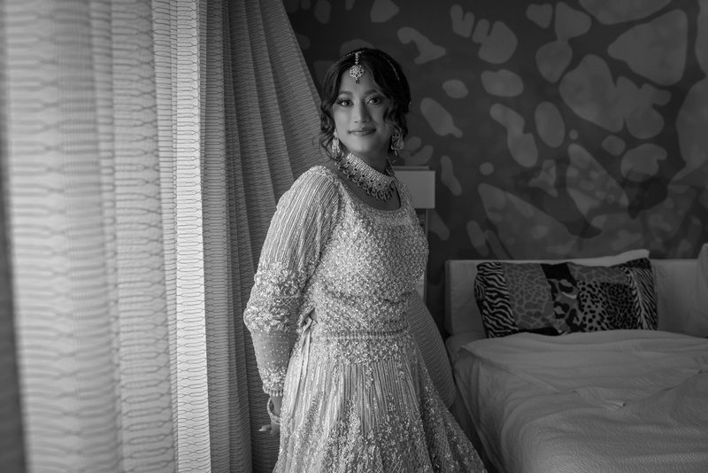 A bride in an ornate traditional gown and jewelry stands calmly in a hotel room with light filtering through curtains.