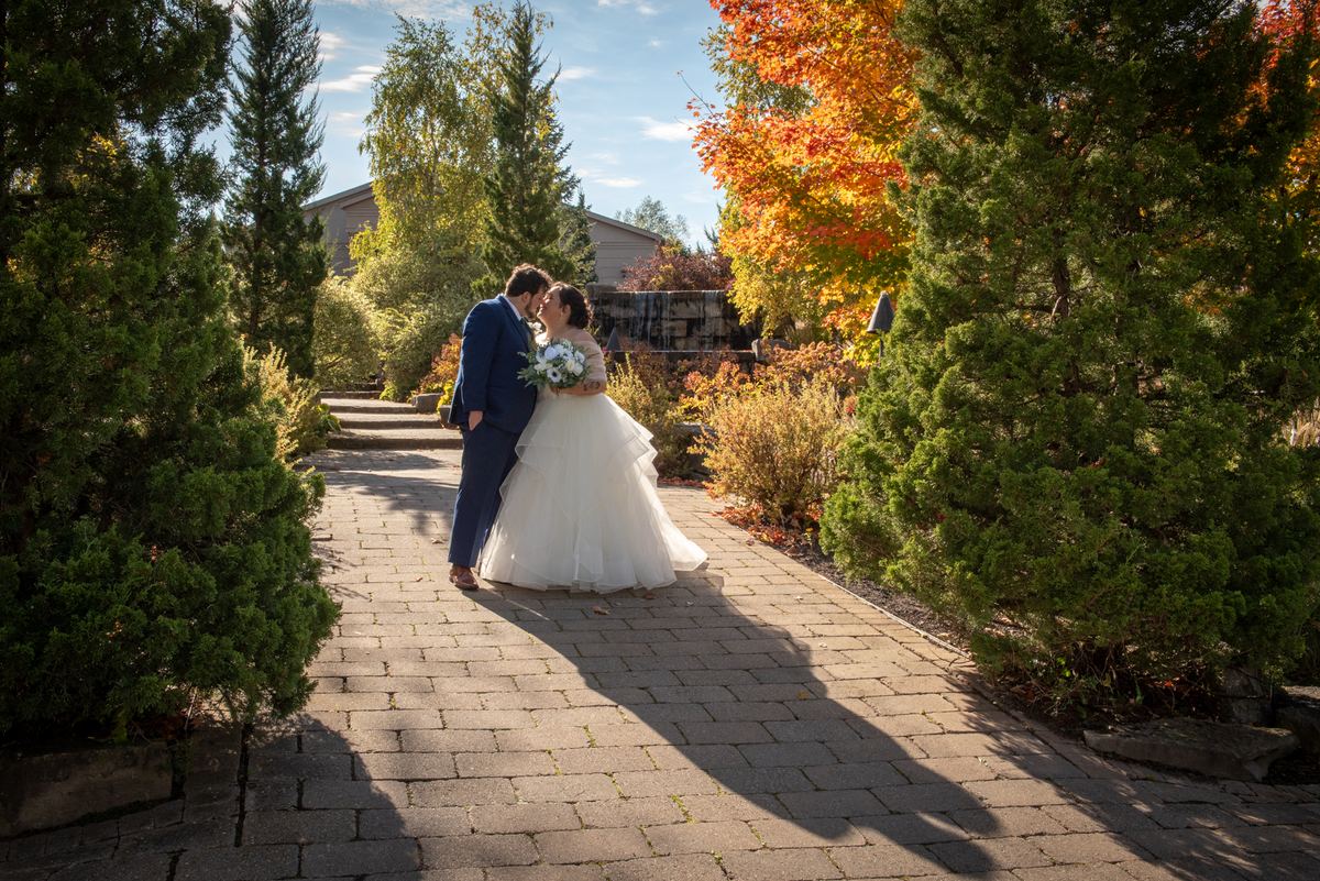A bride and groom kiss on a stone pathway surrounded by autumn leaves and evergreen trees under golden sunlight.