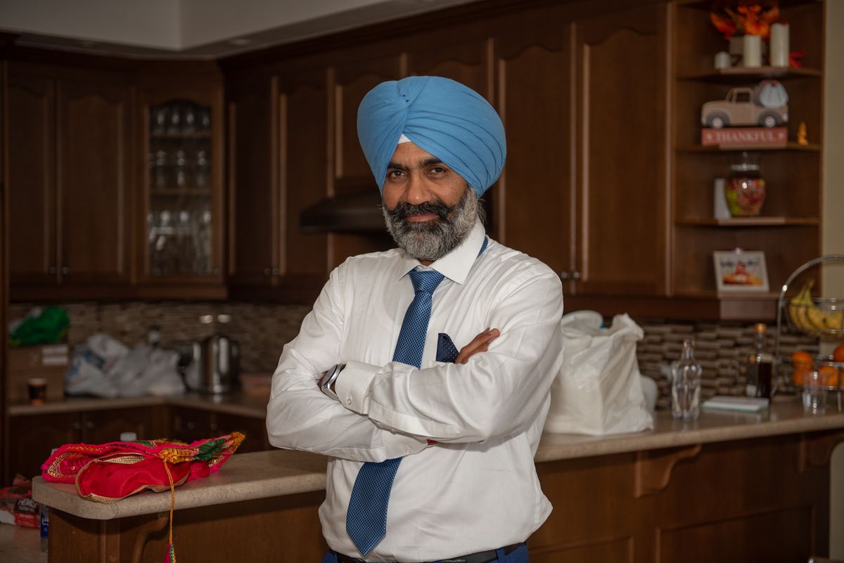 A man in formal wear and a blue turban stands confidently in a warm kitchen with wooden cabinets and colorful textiles.