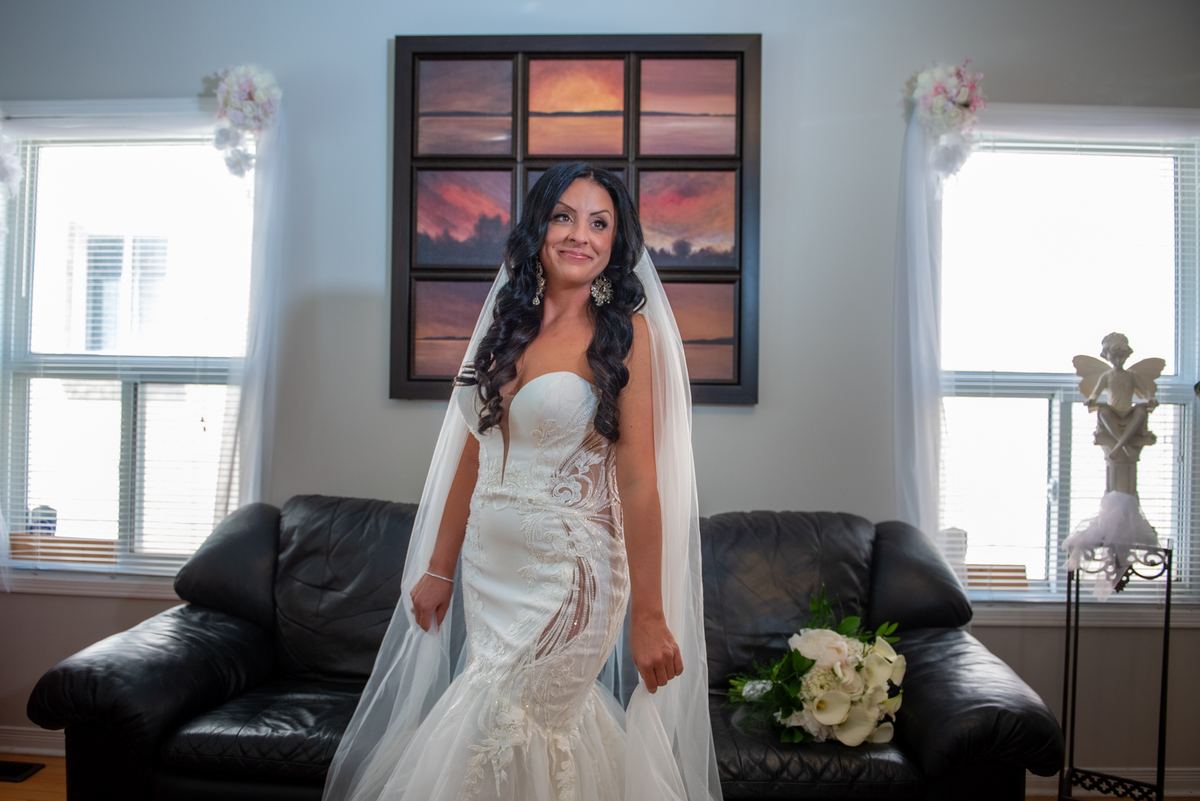 A bride in a white mermaid gown stands smiling with her veil down, bouquet on the sofa beside her in a decorated living room.