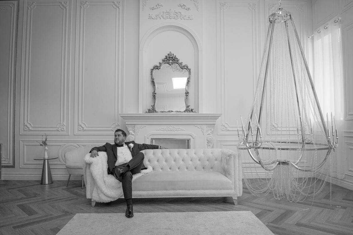 A groom in formal attire relaxes on an ornate white sofa in a grand, Baroque-style room with a cascading chandelier.