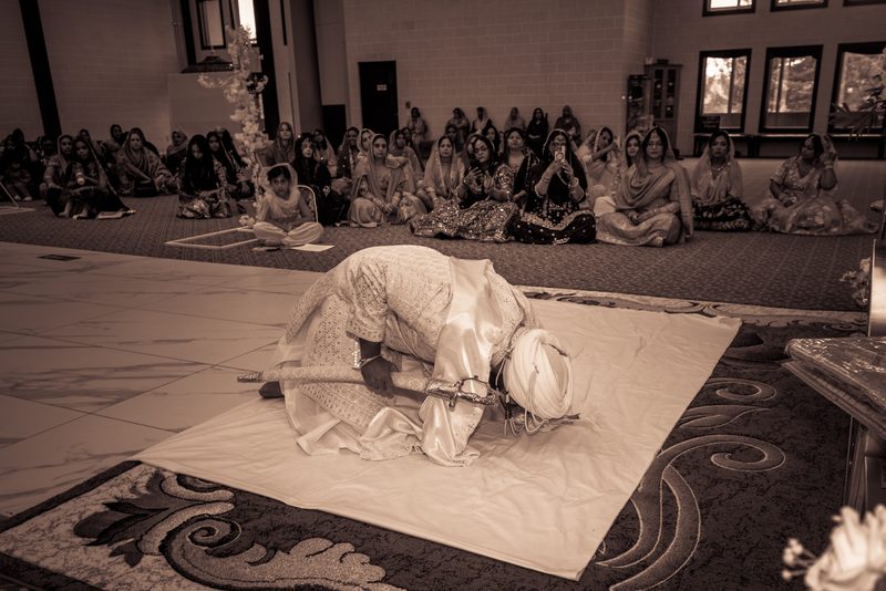A groom in Sikh attire kneels with a ceremonial kirpan as women in colorful dresses observe the solemn moment.