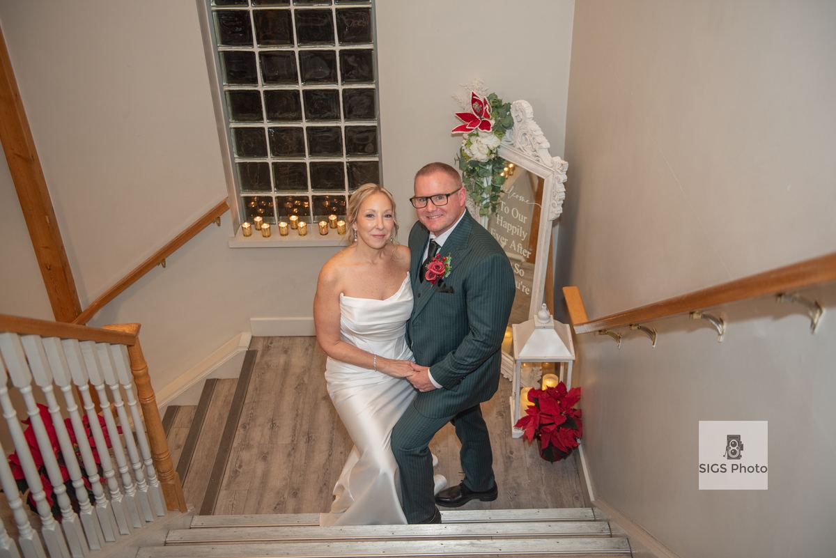 A newlywed couple in formal attire poses on a wooden staircase, surrounded by candles and poinsettias.
