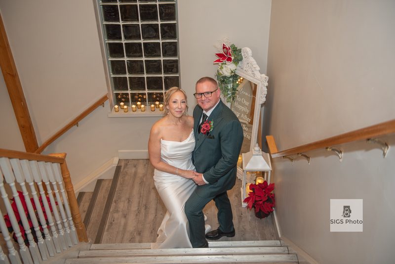 A newlywed couple in formal attire poses on a wooden staircase, surrounded by candles and poinsettias.