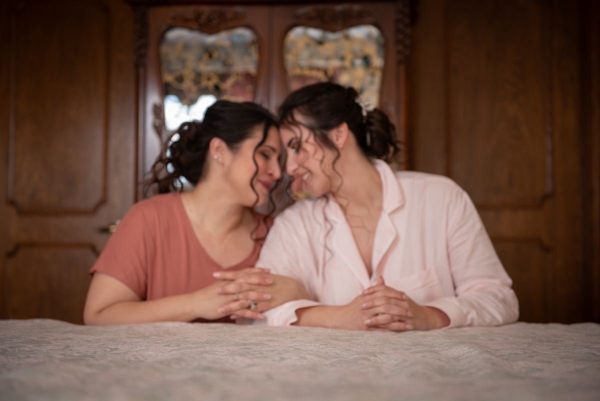 Two women sit closely on a bed, foreheads touching and smiling warmly, hands clasped, in soft light.