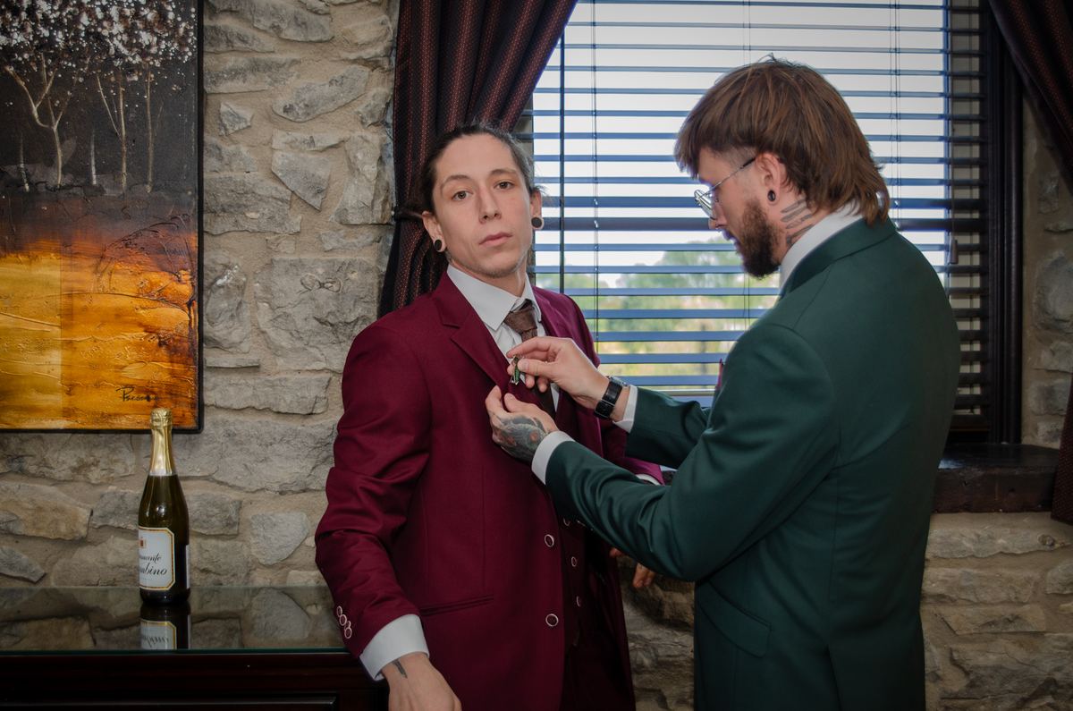A man adjusts another's lapel pin in a warmly lit room with stone walls and horizontal blinds, near a bottle of champagne.