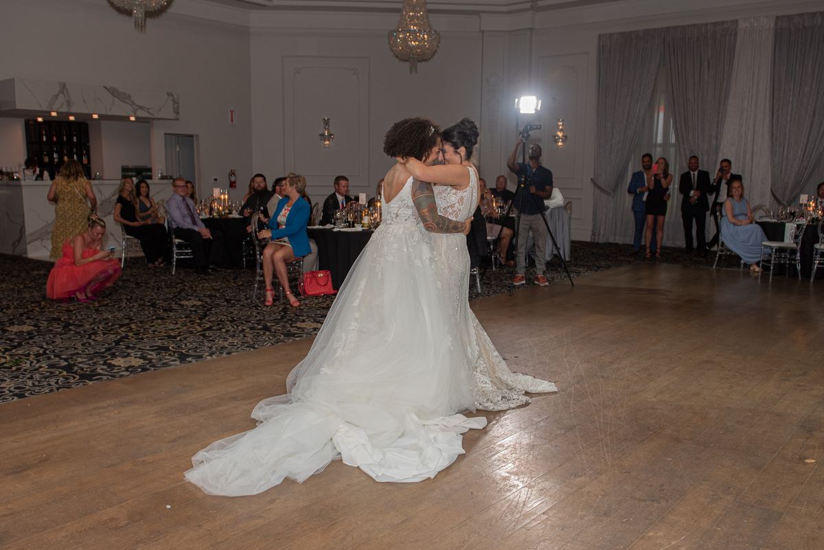 Two brides dance closely in a warmly lit ballroom, surrounded by guests and soft lighting from a chandelier.