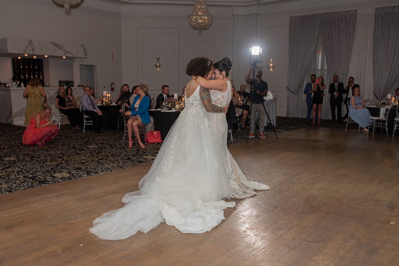 Two brides dance closely in a warmly lit ballroom, surrounded by guests and soft lighting from a chandelier.