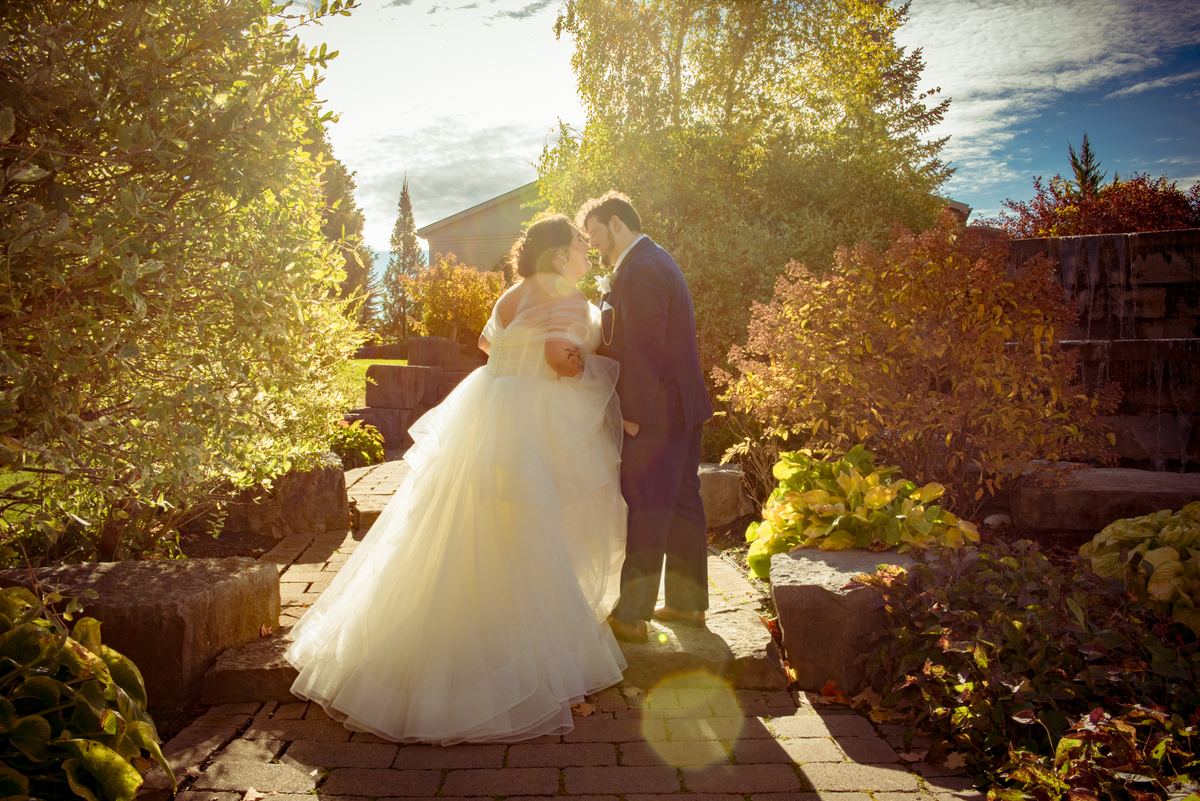 A bride and groom stand close on a stone path, gazing at each other with sunlight filtering through autumn leaves.