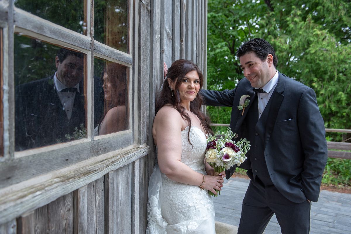 A bride in white lace and groom in black tuxedo smile beside a wooden window with their reflections visible.