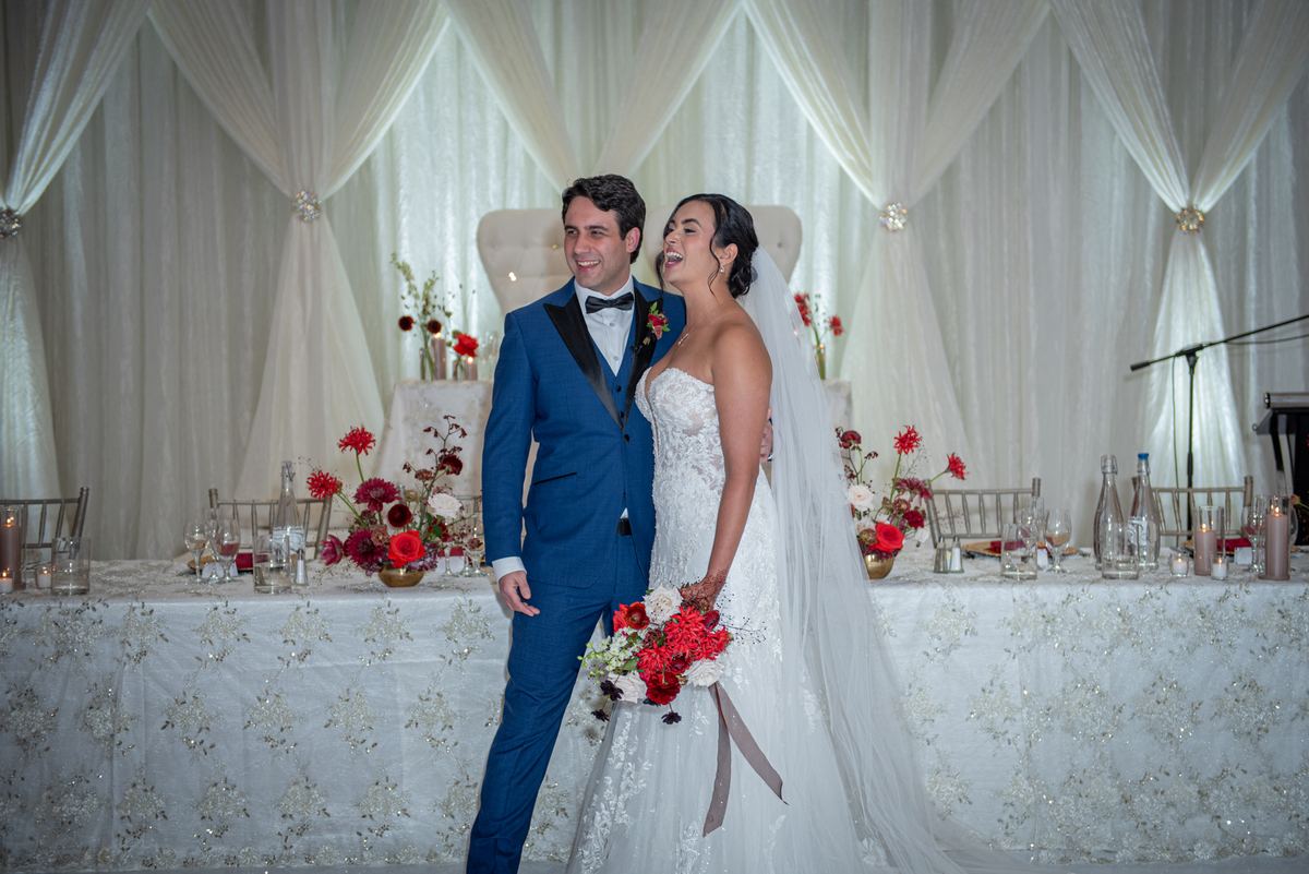 A smiling bride and groom laugh at each other during their reception, lit by soft light through white drapes.