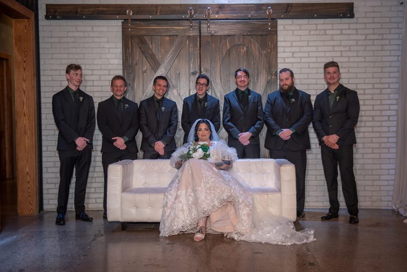 A bride sits with her groom and six groomsmen in dark suits for a wedding portrait against a white brick wall.
