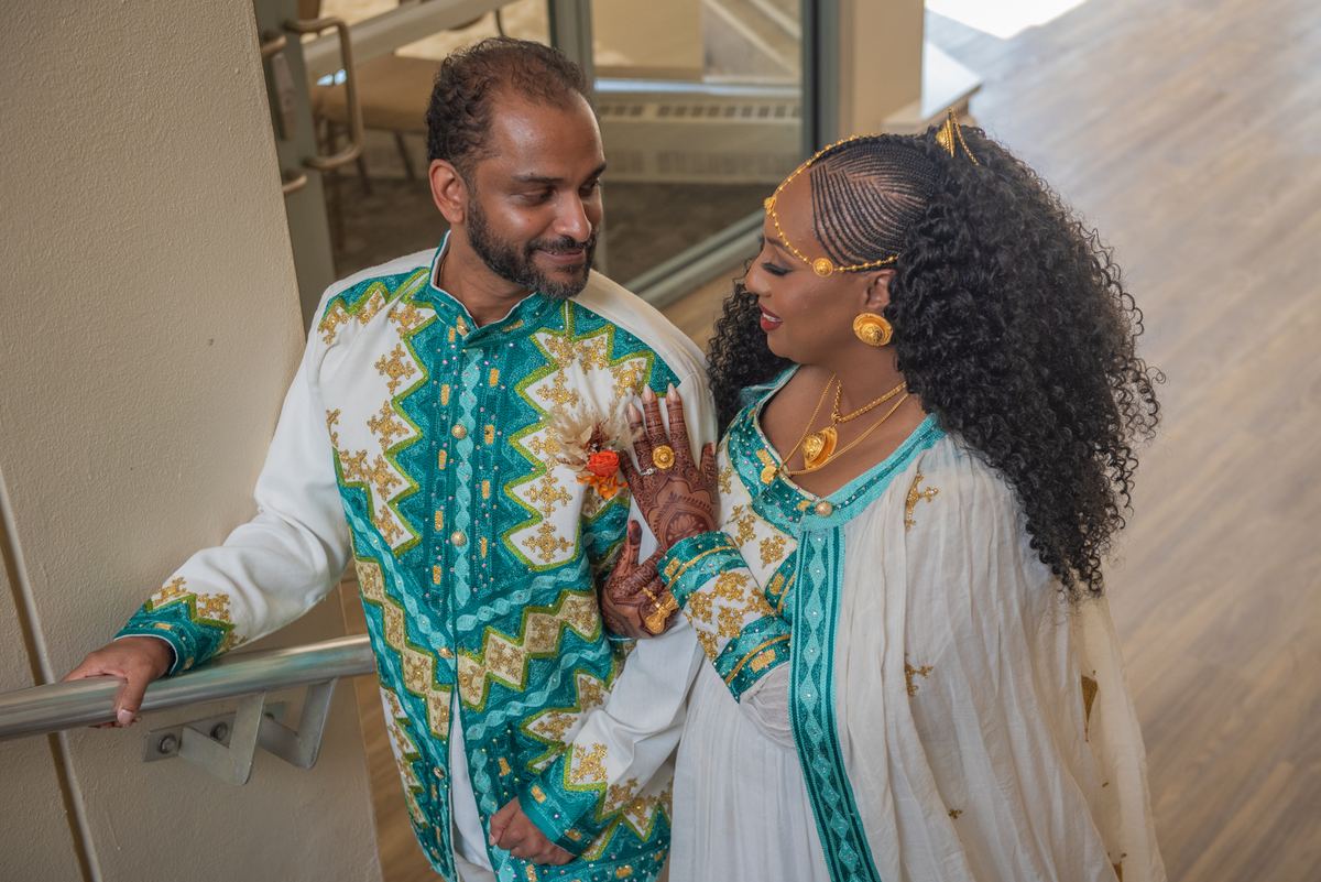 A couple in traditional attire stands closely on a staircase, gazing affectionately at each other under soft light.