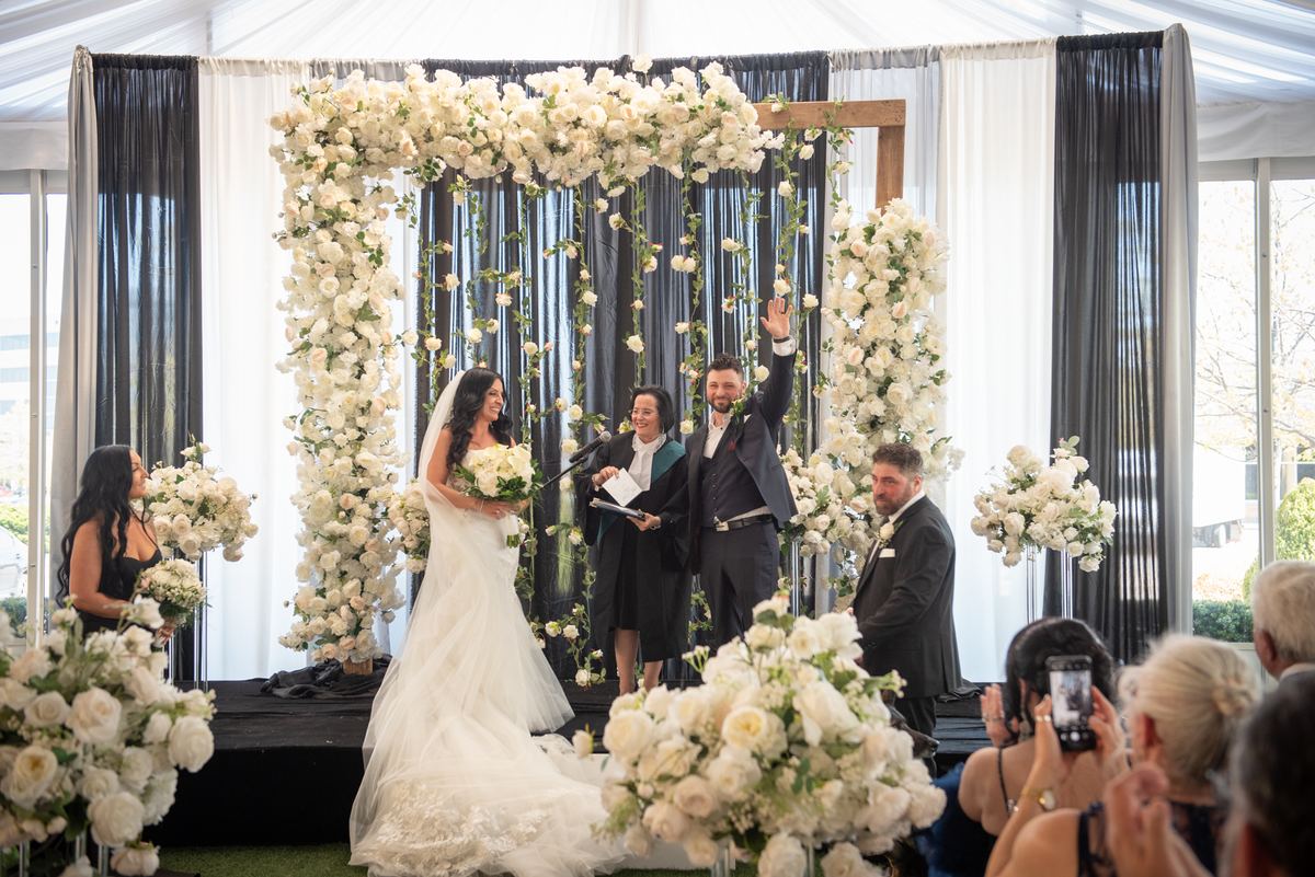 Bride and groom stand under a floral arch of white roses in a tent, guests capturing the moment on phones.