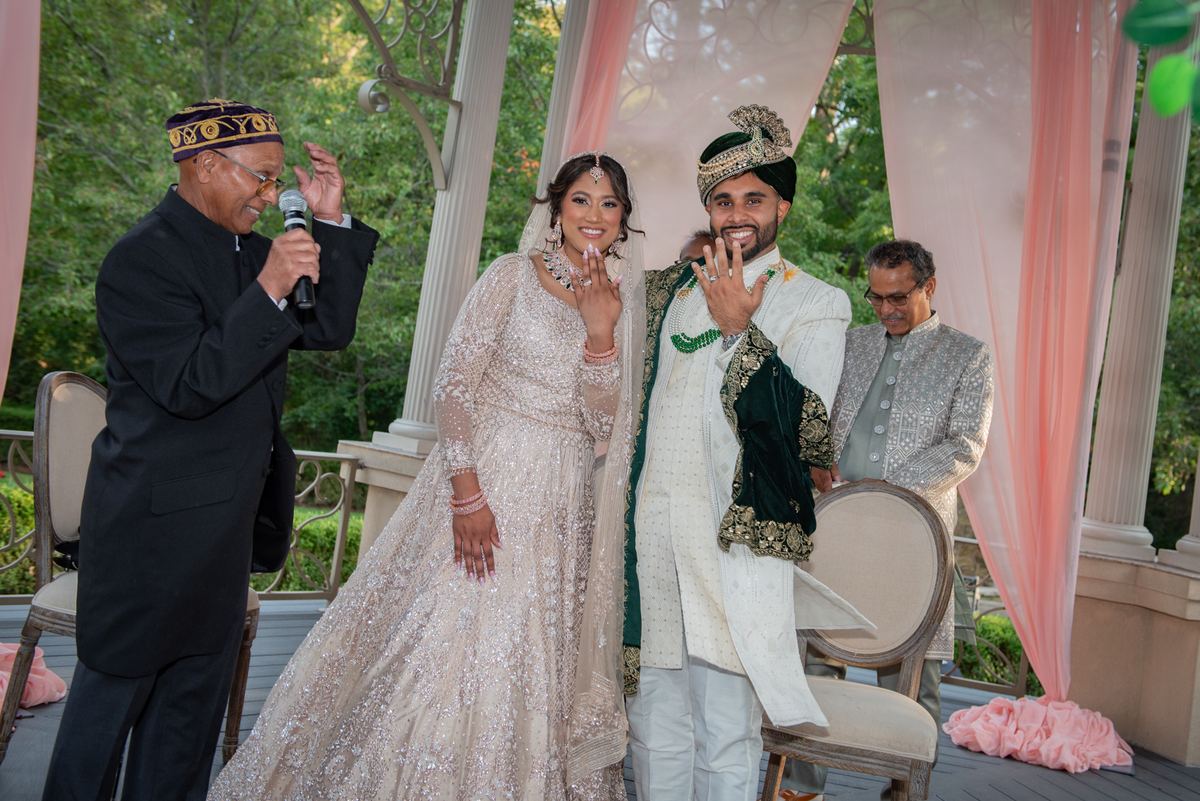 Bride and groom in traditional South Asian attire exchange rings under a decorated canopy, with guests.