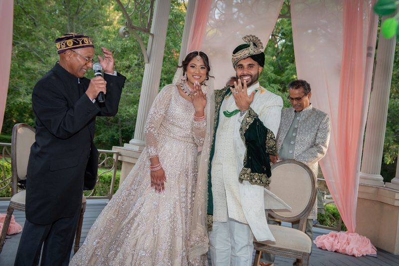 Bride and groom in traditional South Asian attire exchange rings under a decorated canopy, with guests.