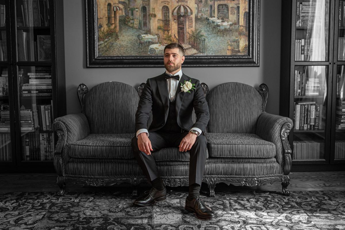 A groom in a tuxedo sits on an ornate sofa, looking at the camera, with natural light from a window illuminating the room.