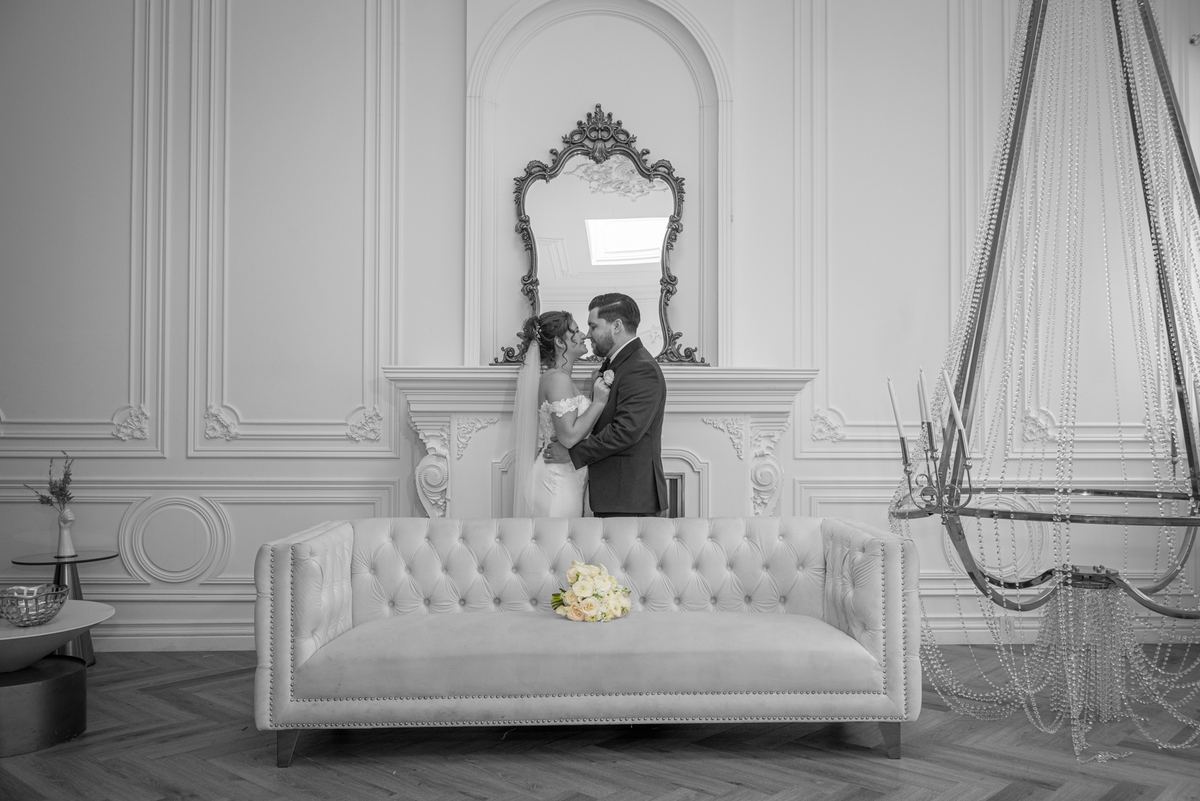 A bride and groom kiss in front of an ornate mirror surrounded by white decor and soft natural light.