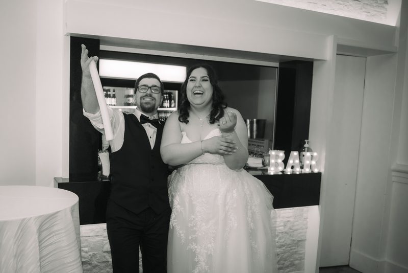 A laughing bride claps as her groom holds up a rolled paper beside a glowing BAR sign at their reception, in black and white.