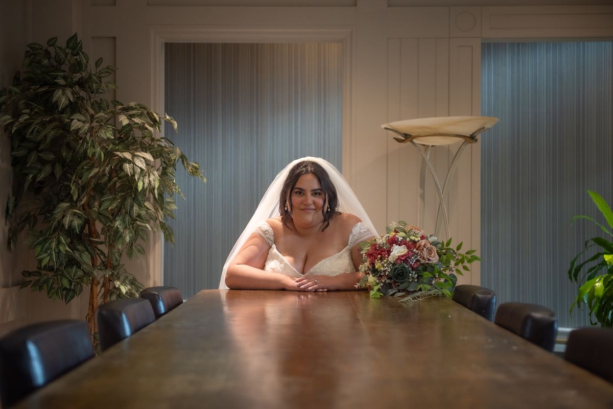 A bride in a white gown and veil sits at a polished wooden conference table, smiling warmly at the camera.