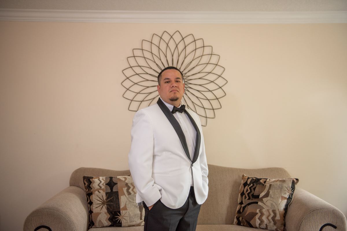 A man in a white tuxedo poses confidently in an elegantly decorated room with a geometric wall sculpture behind him.
