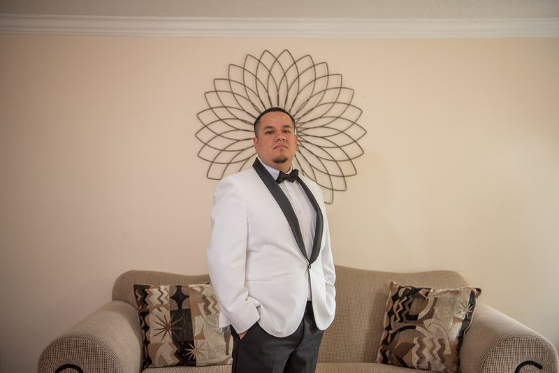 A man in a white tuxedo poses confidently in an elegantly decorated room with a geometric wall sculpture behind him.